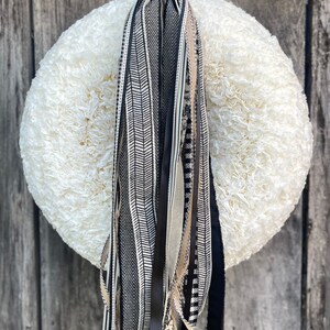 K&ouml;nnte beinhalten: Ein Kranz mit einem Durchmesser von 66 cm und einer cremefarbenen, flauschigen Basis. Dekorative B&auml;nder in Schwarz, Wei&szlig; und Beige, mit Mustern wie Fischgr&auml;ten und Streifen, h&auml;ngen vom Kranz. Der Kranz ist vor einem verwitterten Holzgrund dargestellt.