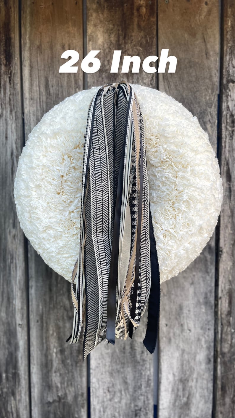 K&ouml;nnte beinhalten: Ein Kranz mit einem Durchmesser von 66 cm und einer cremefarbenen, flauschigen Basis. Dekorative B&auml;nder in Schwarz, Wei&szlig; und Beige, mit Mustern wie Fischgr&auml;ten und Streifen, h&auml;ngen vom Kranz. Der Kranz ist vor einem verwitterten Holzgrund dargestellt.