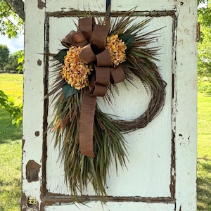 May include: A rustic wreath with a brown ribbon and faux flowers is displayed on a distressed white door. The wreath features brown and green foliage, with a large brown bow and clusters of orange-toned flowers. The door has visible wear and tear.