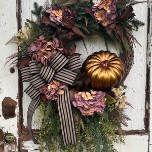 May include: A decorative wreath featuring a metallic bronze pumpkin, clusters of dried purple and peach hydrangea flowers, green pine needles, and brown foliage. A large bow made of black and beige striped ribbon is attached to the side.