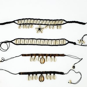 Op de afbeelding: Vier zwarte koordarmbanden met witte schelp kralen en bedels. De armbanden hebben verschillende stijlen en lengtes. Sommige hebben een enkele schelp bedel, terwijl andere meerdere schelp bedels hebben.