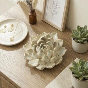 May include: A ceramic succulent sculpture in a cream color with subtle blue-grey speckles, shaped like a blooming flower with layered petals. The sculpture is displayed alongside a white plate with gold earrings, a small brown glass vase, a framed line drawing, and two potted green succulents.