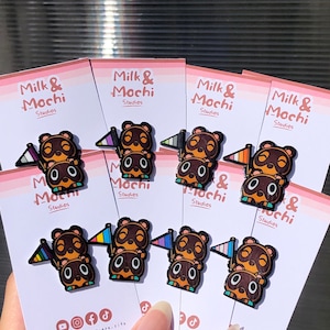 Könnte beinhalten: Sechs Emaille-Pins mit einer Comicfigur aus dem Videospiel Animal Crossing. Die Figur ist ein brauner Bär mit einer weißen Schnauze und einer schwarzen Nase. Die Figur trägt ein rotes Hemd und eine blaue Mütze. Die Pins sind in einer Reihe angeordnet, wobei jeder Pin eine andere Flagge zeigt. Die Flaggen sind Regenbogen, Transgender, Bisexuell, Lesbisch, Pansexuell und Asexuell. Die Pins befinden sich alle auf einem rosa Hintergrund mit dem Text "Milk & Mochi Studios" auf jeder Karte.