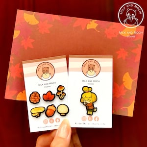 May include: Two sets of enamel pins featuring a cartoon dog and mushroom designs. The pins are on white cards with the text "Milk and Mochi Studios" and social media icons.