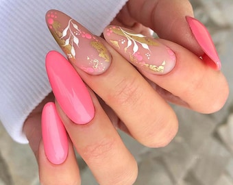 May include: Close-up of a hand with pink and nude almond-shaped nails. The nude nails have a gold leaf design with white floral accents.