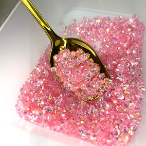 Könnte beinhalten: Ein goldener Löffel schöpft rosa schimmernde Kristallperlen aus einem weißen Behälter. Die Perlen sind klein und haben eine glitzernde Oberfläche.