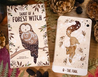 以下が含まれることがあります： Tarot of a Forest Witchのデッキボックスと「0 - The Fool」と書かれたカード1枚。箱には白い花柄のフクロウのイラストが描かれています。カードには、白い渦巻き線と星に囲まれたキツネが描かれています。