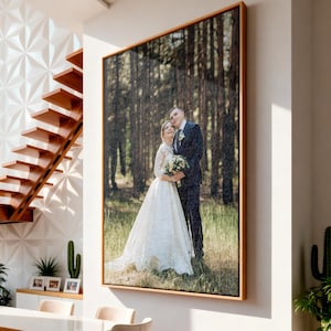 May include: A large rectangular framed wall art piece featuring a photograph of a couple in wedding attire standing in a pine forest. The artwork is displayed on a white wall above a dining table set with chairs in a bright, modern interior space.