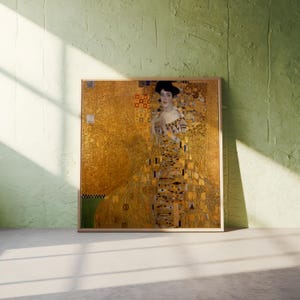May include: A framed print of a Gustav Klimt painting, featuring a woman in an ornate gold dress. The artwork has a gold background with geometric patterns and a touch of green. The frame is a light brown color.