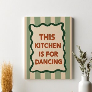 May include: A rectangular canvas art piece with a green and beige striped background. The artwork features a brown framed text that reads "THIS KITCHEN IS FOR DANCING". A white vase with green stems is on the right.