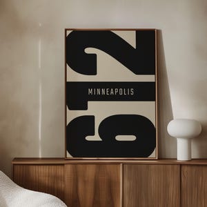 May include: A framed art print featuring the city name "Minneapolis" in gold lettering on a black horizontal band. Large black numbers are above and below the band on a beige background. The artwork is displayed on a wooden cabinet.