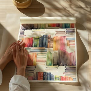 May include: Watercolor print featuring a repeating pattern of colorful books. The books are arranged in stacks and rows, with a variety of colors including red, blue, green, and orange. The print is on a roll of paper, with a light-colored background.