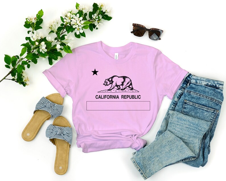 May include: A pink t-shirt with a black graphic of the California bear and star, with the text "CALIFORNIA REPUBLIC" underneath. The shirt is laid out on a white surface with a pair of blue jeans, a pair of striped sandals, and a pair of sunglasses.