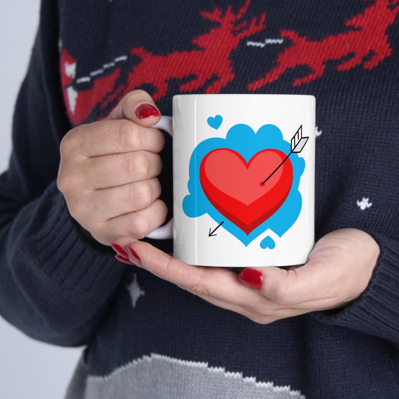 May include: White ceramic mug with a blue cloud and a red heart pierced by an arrow. The heart is 3D and has a red outline. There are small white hearts around the cloud.