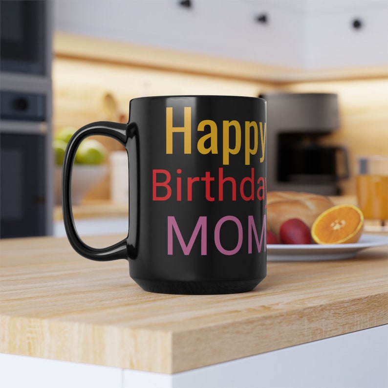 May include: Black coffee mug with a white handle. The mug has the text "Happy Birthday MOM" in colorful letters.