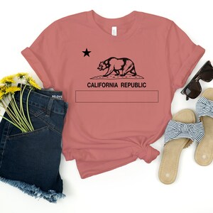 May include: A dusty rose t-shirt with a black graphic of the California Republic flag featuring a bear and a star. The text "CALIFORNIA REPUBLIC" is printed below the graphic.