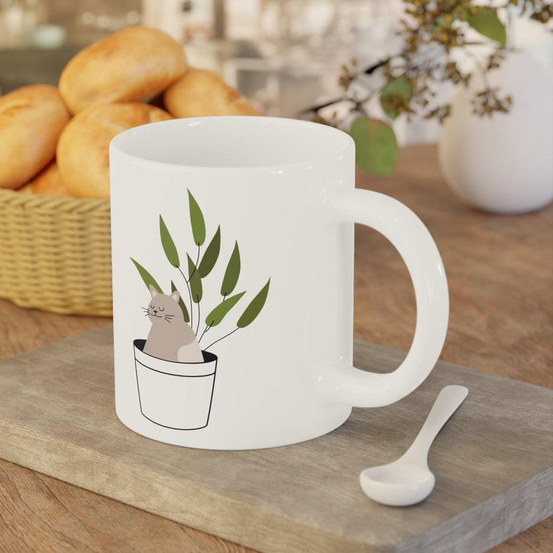 May include: White ceramic mug with a graphic of a cat sitting in a pot with green leaves. The cat is light brown and has closed eyes.