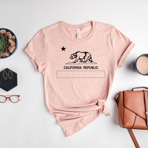 May include: A pink t-shirt with a black graphic of the California bear and the text "California Republic". The shirt is laid flat on a white surface with a potted succulent, a pair of glasses, a small black hexagonal box with earrings, a brown leather purse, and a blue mug with coffee.