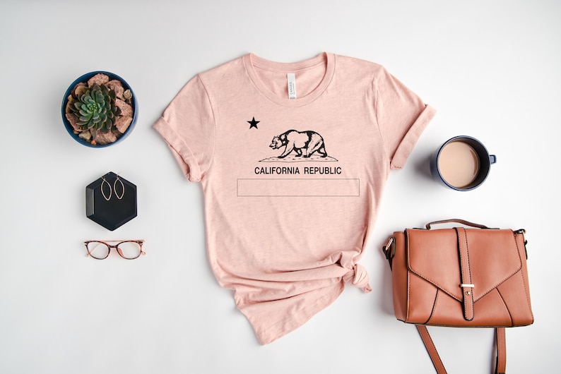 May include: A pink t-shirt with a black graphic of the California bear and the text "California Republic". The shirt is laid flat on a white surface with a potted succulent, a pair of glasses, a small black hexagonal box with earrings, a brown leather purse, and a blue mug with coffee.
