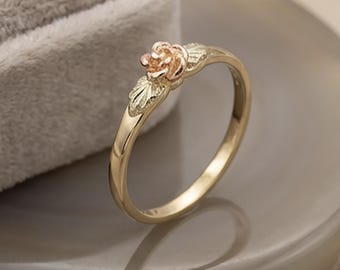 May include: A gold-colored ring with a rose and leaf design. The ring features a small, detailed rose at the top and delicate leaves on either side. The ring is displayed on a neutral-toned surface.