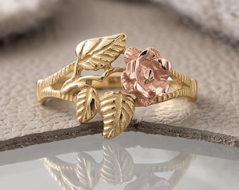 May include: A gold ring featuring a rose gold flower and yellow gold leaves. The band has a textured, branch-like design that supports the floral motif. The metal surfaces are polished, reflecting light to highlight the intricate details of the petals.