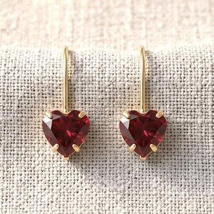 May include: A pair of gold drop earrings featuring faceted, heart-shaped deep red gemstones. Each stone is secured by three small gold prongs, attached to a slender gold wire hook that extends upwards from the top of the heart setting.
