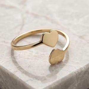 May include: A gold-toned open ring featuring two smooth, polished heart-shaped ends that face each other. The band is thin and circular, creating a delicate and minimalist appearance with a reflective metallic finish on the heart motifs.