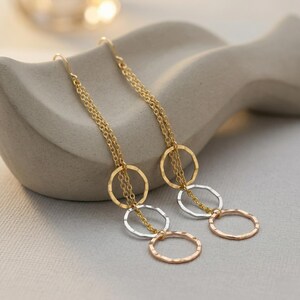 Puede incluir: Pendientes colgantes dorados, plateados y oro rosa. Cada pendiente presenta tres anillos texturizados, unidos por cadenas. El anillo superior es dorado, el central plateado y el inferior oro rosa. Se muestran sobre una superficie de color neutro.
