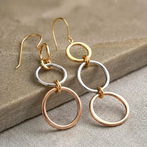 Puede incluir: Pendientes colgantes en oro, plata y oro rosa. Cada pendiente presenta tres aros conectados en diferentes tonos metálicos. El aro superior es dorado, el central plateado y el inferior oro rosa. Cierre de gancho.