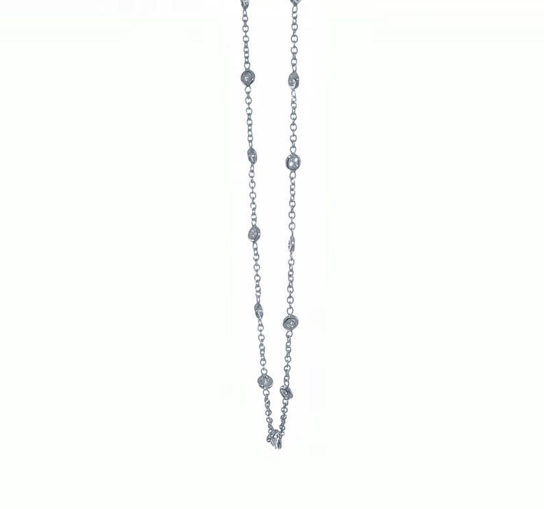 May include: A delicate silver necklace featuring multiple round, clear gemstones evenly spaced along the chain. The necklace is displayed against a plain white background, highlighting the sparkle of the gemstones.