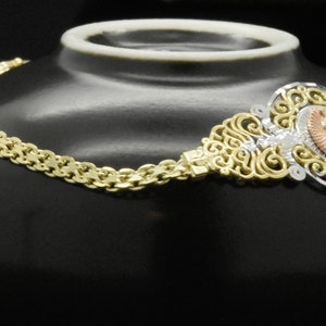 May include: A gold chain bracelet with a filigree design and a rose gold oval pendant. The pendant has a floral design and is set with clear stones.