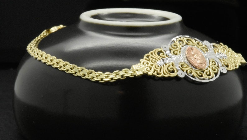 May include: A gold chain bracelet with a filigree design and a rose gold oval pendant. The pendant has a floral design and is set with clear stones.