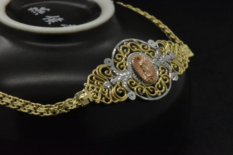 May include: A gold chain necklace with a filigree pendant featuring a religious image in the center. The pendant is made of gold, silver, and rose gold.