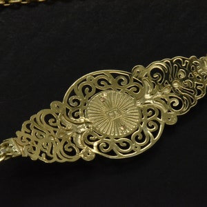 May include: A gold chain necklace with a decorative pendant featuring a circular design with intricate filigree details. The pendant has a raised center with a sunburst pattern.