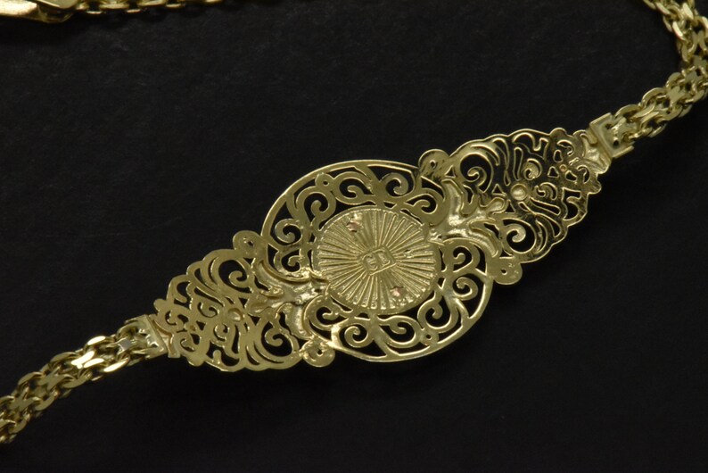 May include: A gold chain necklace with a decorative pendant featuring a circular design with intricate filigree details. The pendant has a raised center with a sunburst pattern.