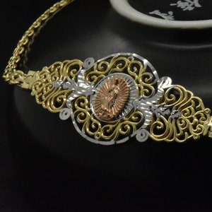 May include: A gold and silver filigree bracelet with a rose gold medallion featuring a religious figure. The bracelet is on a black background.