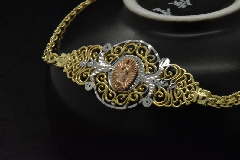 May include: A gold and silver filigree bracelet with a rose gold medallion featuring a religious figure. The bracelet is on a black background.