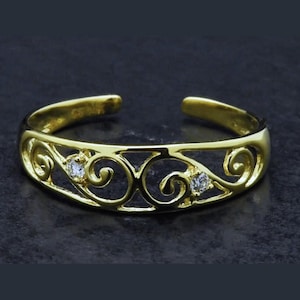 May include: A gold-colored toe ring with an open design. The ring features a filigree pattern with two small, clear gemstones. The band is adjustable and has a polished finish, set against a dark background.