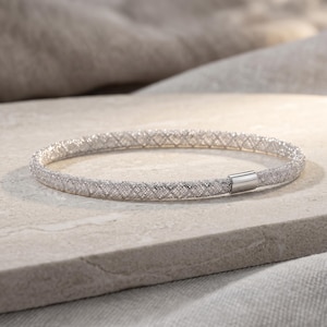 May include: A silver-toned bangle bracelet featuring a fine, textured mesh design that creates a sparkling, grid-like pattern across its surface. The circular bracelet is finished with a smooth, polished silver-colored metal clasp connector.