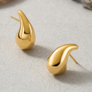 Op de afbeelding: Twee goudkleurige druppelvormige oorbellen. Elke oorbel heeft een glad, reflecterend oppervlak en een pin voor doorboorde oren. De oorbellen worden weergegeven op een lichtgrijze ondergrond, met zachte schaduwen.