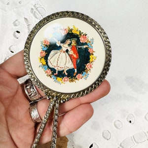 May include: A vintage hand mirror with a silver-toned frame and a floral design. The mirror features a colourful image of a couple dancing in a circle of flowers.