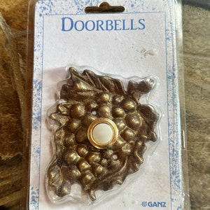 May include: A gold-toned doorbell button with a grapevine design. The button is packaged in a clear plastic clamshell with a white card that reads "Doorbells" and "GANZ".