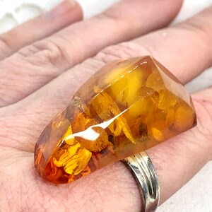 May include: A large, amber-colored ring with a unique, faceted design. The ring features inclusions of fossilized plant material, giving it a natural, organic look.
