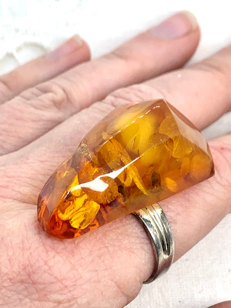 May include: A large, amber-colored ring with a unique, faceted design. The ring features inclusions of fossilized plant material, giving it a natural, organic look.