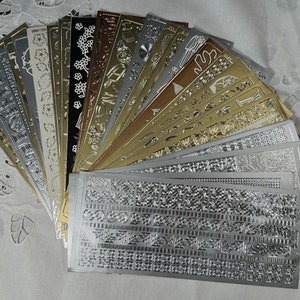 Peut inclure: Une collection d'embellissements adhésifs dorés et argentés, présentant divers motifs et designs. Les embellissements sont disposés en éventail, les dorés en haut et les argentés en bas.