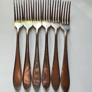 May include: Six vintage silver forks with tines and teardrop-shaped handles. The forks are arranged in a row, showcasing their polished metal finish. The handles have a copper-toned patina, adding a touch of antique charm to the dining utensils.