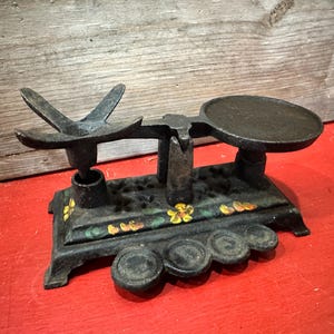 May include: Antique cast iron scale with a decorative bird-shaped counterweight and a round tray. The scale is painted black with floral accents in yellow, orange, and green. The scale is displayed on a red surface.