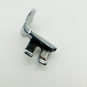 May include: A silver metal sewing machine foot attachment with a curved shape and two prongs.