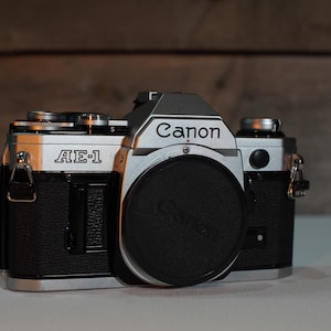 May include: A silver Canon AE-1 film camera with a black lens and a black leather strap. The camera has a silver top plate with the word "Canon" printed in black.