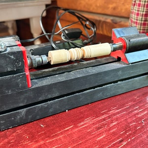 Könnte beinhalten: Nahaufnahme einer kleinen Holzdrehmaschine mit einem Stück Holz, das gedreht wird. Die Drehmaschine ist schwarz mit roten Akzenten und steht auf einem Holztisch.